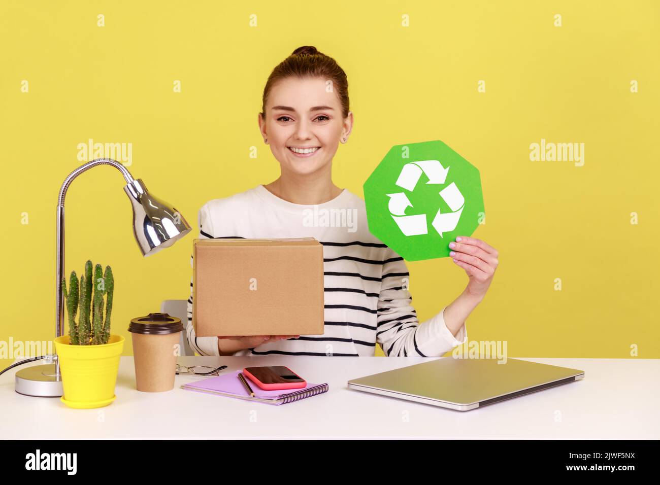 Verantwortungsbewusste selbstbewusste Frau mit grünem Recycling-Schild in der Hand und Pappkarton, sitzend am Arbeitsplatz mit Laptop. Studio-Innenaufnahme isoliert auf gelbem Hintergrund. Stockfoto