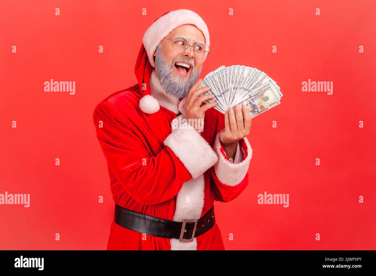 Porträt eines älteren Mannes mit grauem Bart, der ein weihnachtsmann-Kostüm mit aufgeregter Expression trägt, gieriger Mann mit Dollarfan, der Banknoten ansieht. Innenaufnahme des Studios isoliert auf rotem Hintergrund. Stockfoto