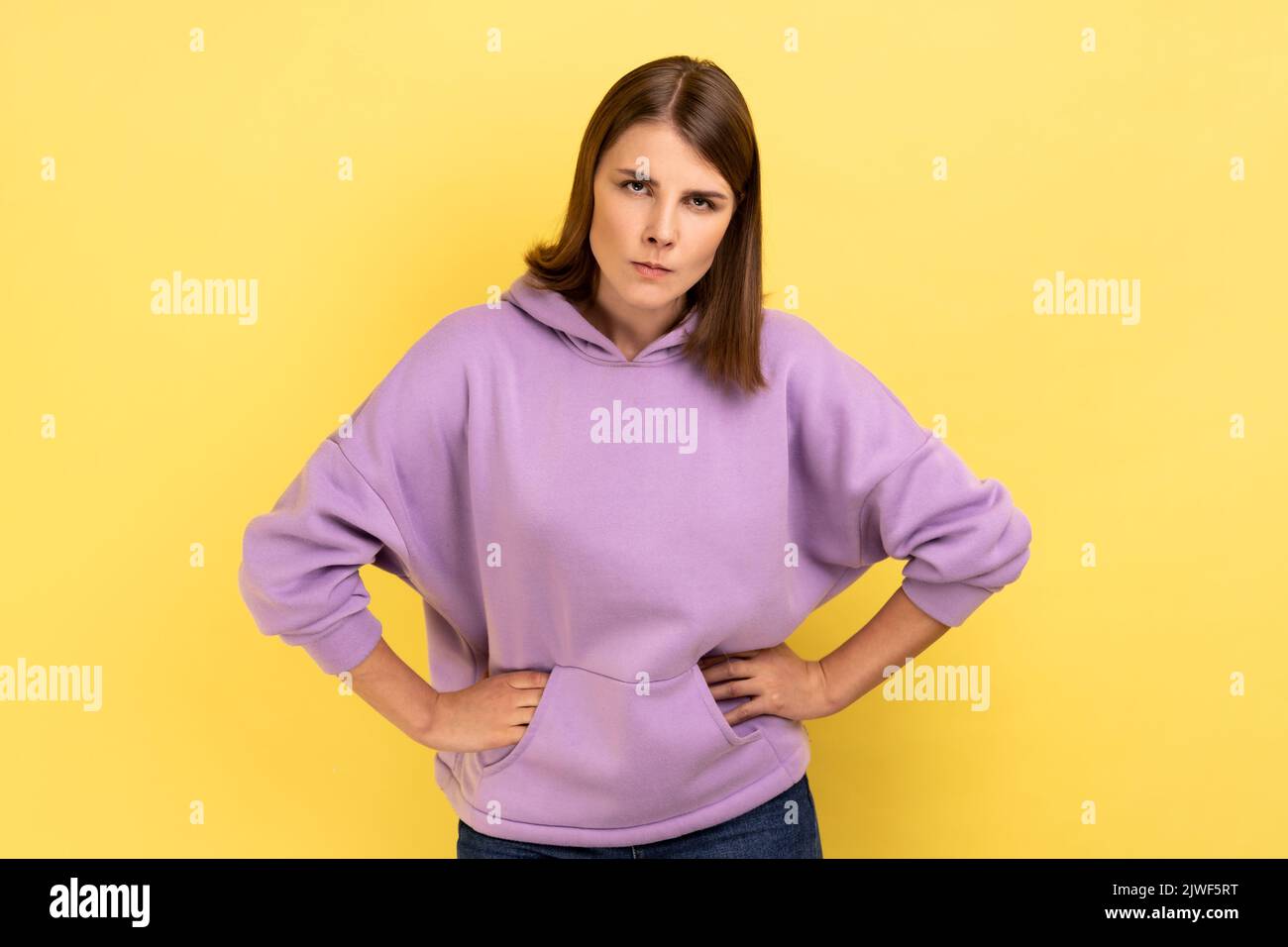 Porträt einer jungen, wütenden, ernsthaften Frau, die mit strengem Negativausdruck, Aggression, Händen an den Hüften und purpurfarbenem Hoodie auf die Kamera schaut. Innenaufnahme des Studios isoliert auf gelbem Hintergrund. Stockfoto