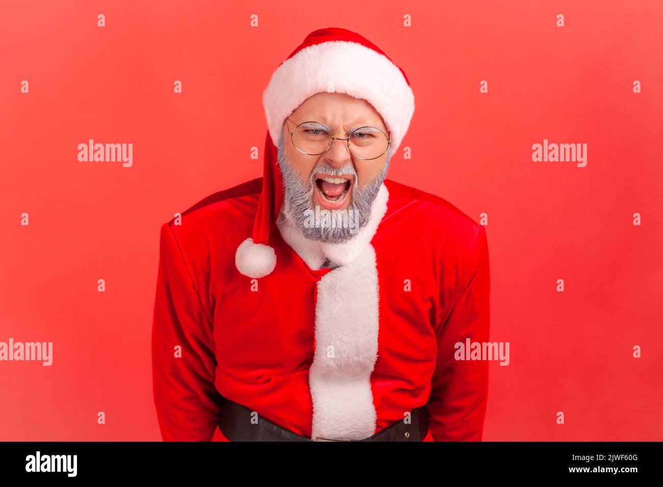 Wütender älterer Mann mit grauem Bart, der das weihnachtsmann Kostüm trägt und laut schreit, mit aggressivem Ausdruck, die Kamera anschaut und schreit. Innenaufnahme des Studios isoliert auf rotem Hintergrund. Stockfoto