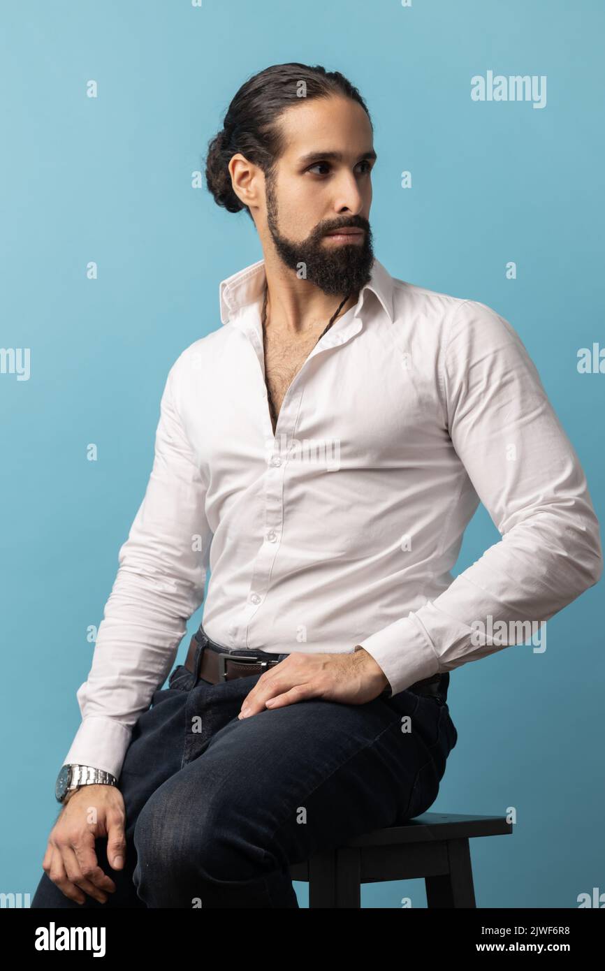 Porträt eines attraktiven Mannes mit Bart und dunklen Haaren in weißem Hemd und schwarzer Hose, sitzend und mit selbstbewusstem Ausdruck wegblickend. Innenaufnahme des Studios isoliert auf blauem Hintergrund Stockfoto