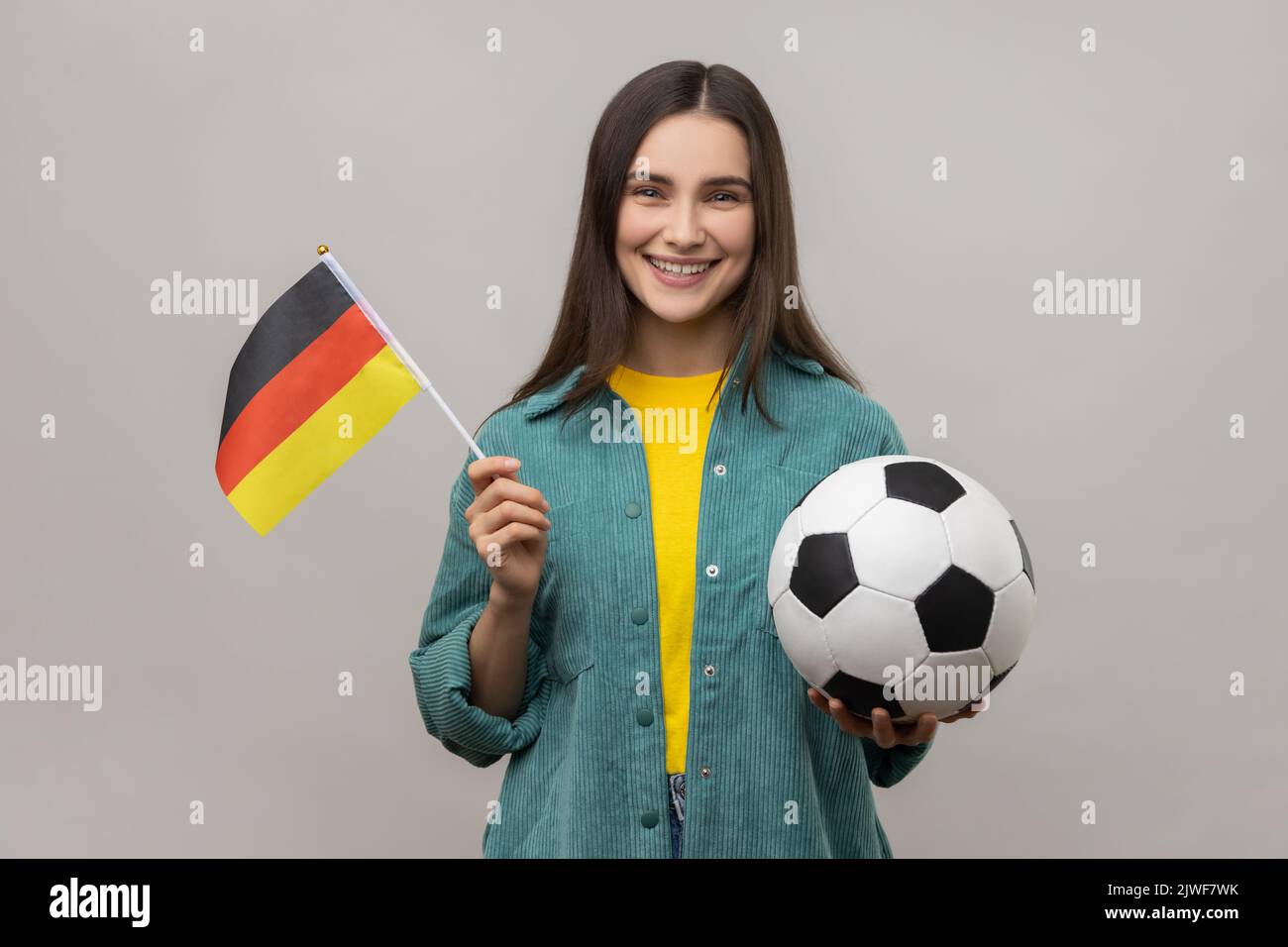 Lächelnde, zufriedene Frau, die die Flagge Deutschlands und des Fußballs in den Händen hält, die das Lieblingsteam in der Meisterschaft unterstützt und eine Jacke im lässigen Stil trägt. Innenaufnahme des Studios isoliert auf grauem Hintergrund. Stockfoto