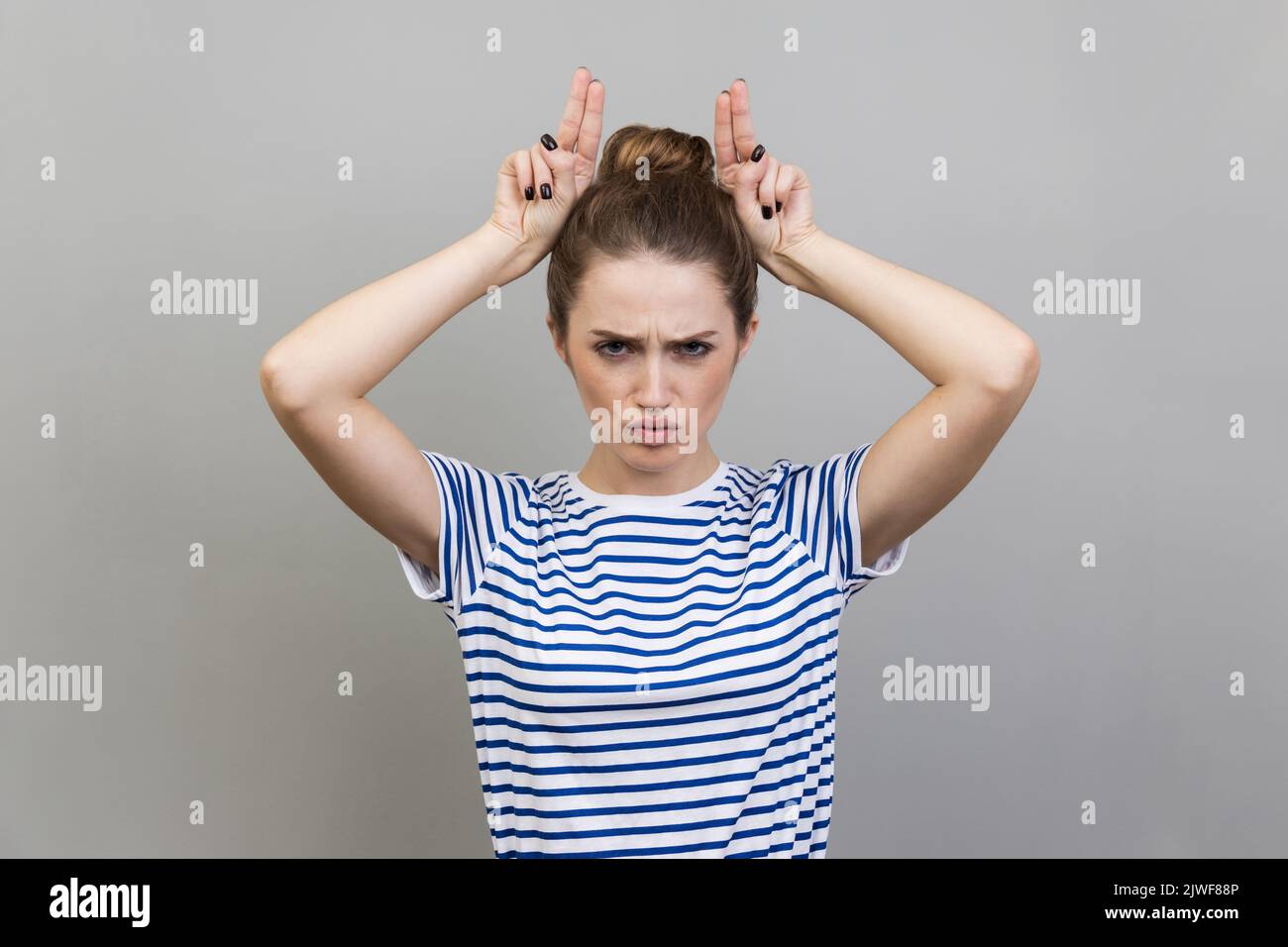 Porträt einer wütenden, tyrannisieren Frau in T-Shirt mit Bullenhörnern mit Fingern auf dem Kopf, Geweih-Geste, bedrohlich mit feindseliger Sicht, angriffbereit. Innenaufnahme des Studios isoliert auf grauem Hintergrund. Stockfoto