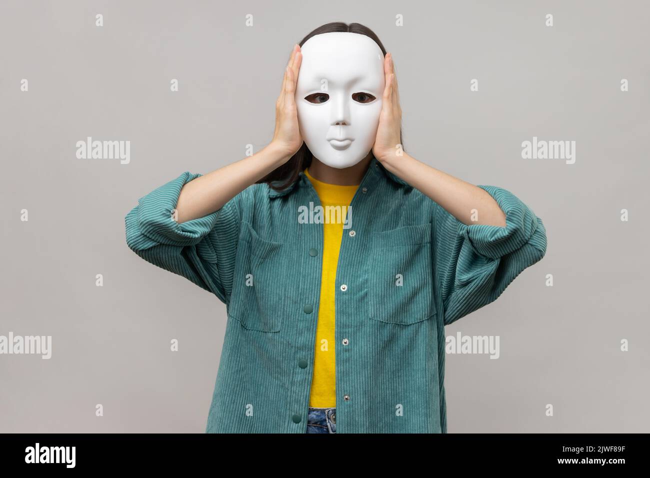 Porträt einer unbekannten Frau mit weißer Maske, stehend bedeckendes Gesicht, Multiple Persönlichkeitsstörung, tragen Jacke im lässigen Stil. Innenaufnahme des Studios isoliert auf grauem Hintergrund. Stockfoto