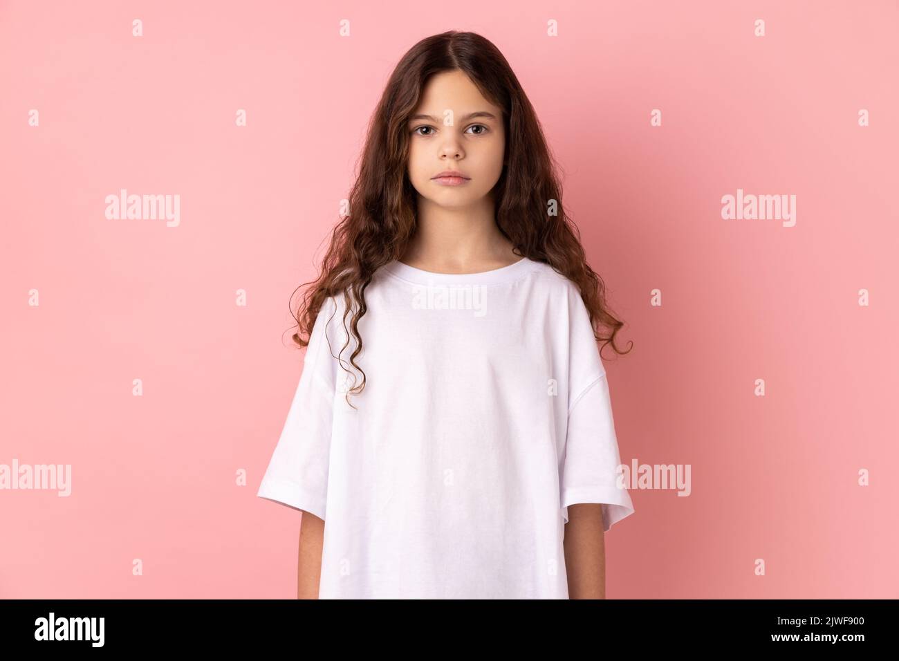Porträt eines ernsthaften strengen kleinen Mädchens, das weißes T-Shirt trägt und die Kamera mit bossiger Ausdruckskraft anschaut, in schlechter Laune. Innenaufnahme des Studios isoliert auf rosa Hintergrund. Stockfoto