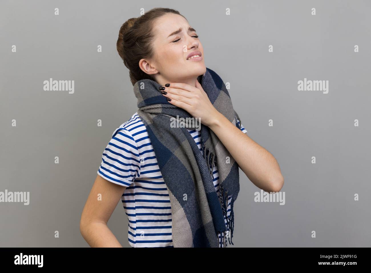Kranke Frau im T-Shirt und in Schal gehüllt, berührt den Hals, leidet an Halsschmerzen, kann nicht gut atmen, würgt und grinst Gesicht, fühlt sich unwohl. Innenaufnahme des Studios isoliert auf grauem Hintergrund. Stockfoto