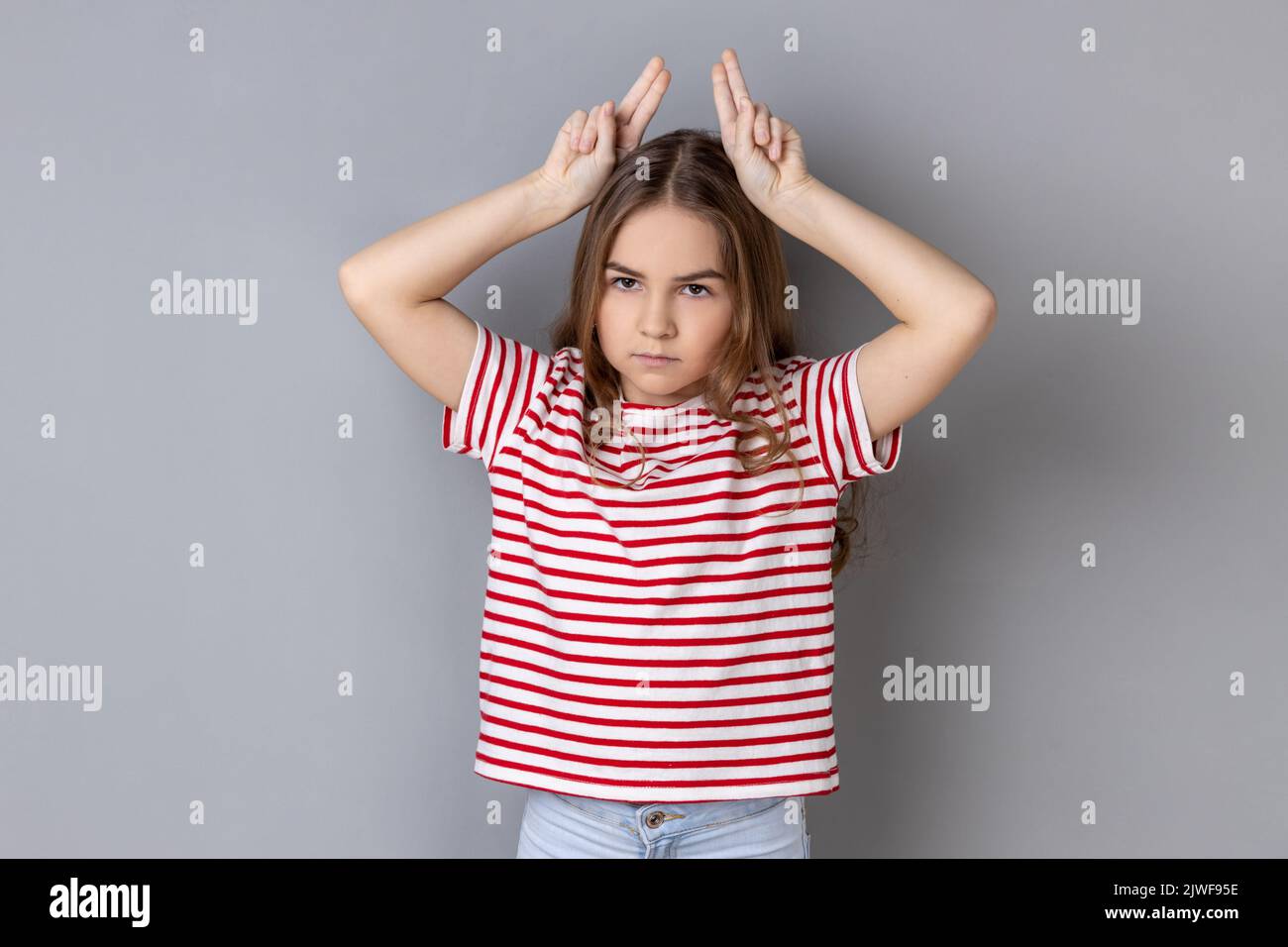 Porträt eines aggressiven, tyrannisierten kleinen Mädchens in einem gestreiften T-Shirt mit Stierhörnern über dem Kopf und Stirnrunzeln wie vor dem Angriff. Innenaufnahme des Studios isoliert auf grauem Hintergrund. Stockfoto
