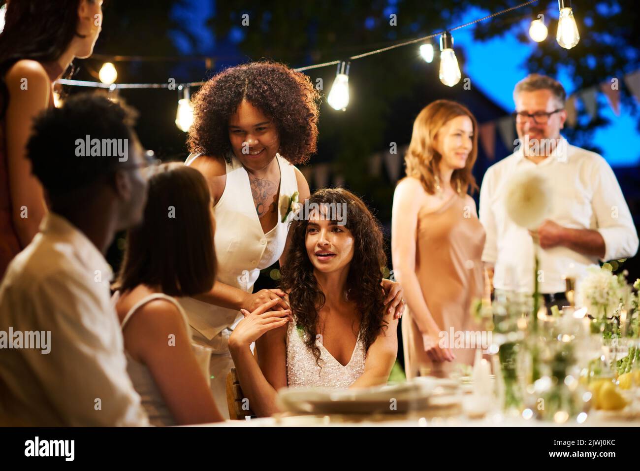 Glückliche interkulturelle lesbische Mädchen in Hochzeitskleidung im Gespräch mit ihren Freunden am gedienten Tisch gegen reifes heterosexuelles Paar auf der Party Stockfoto
