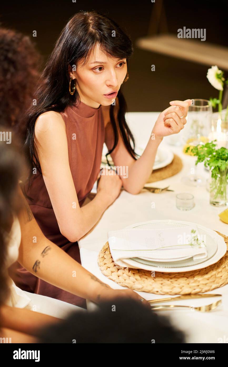 Junge Brünette Frau in elegantem SeidenAbendkleid sitzen am gedienten Tisch während der Hochzeit Fest und im Gespräch mit anderen Gästen Stockfoto