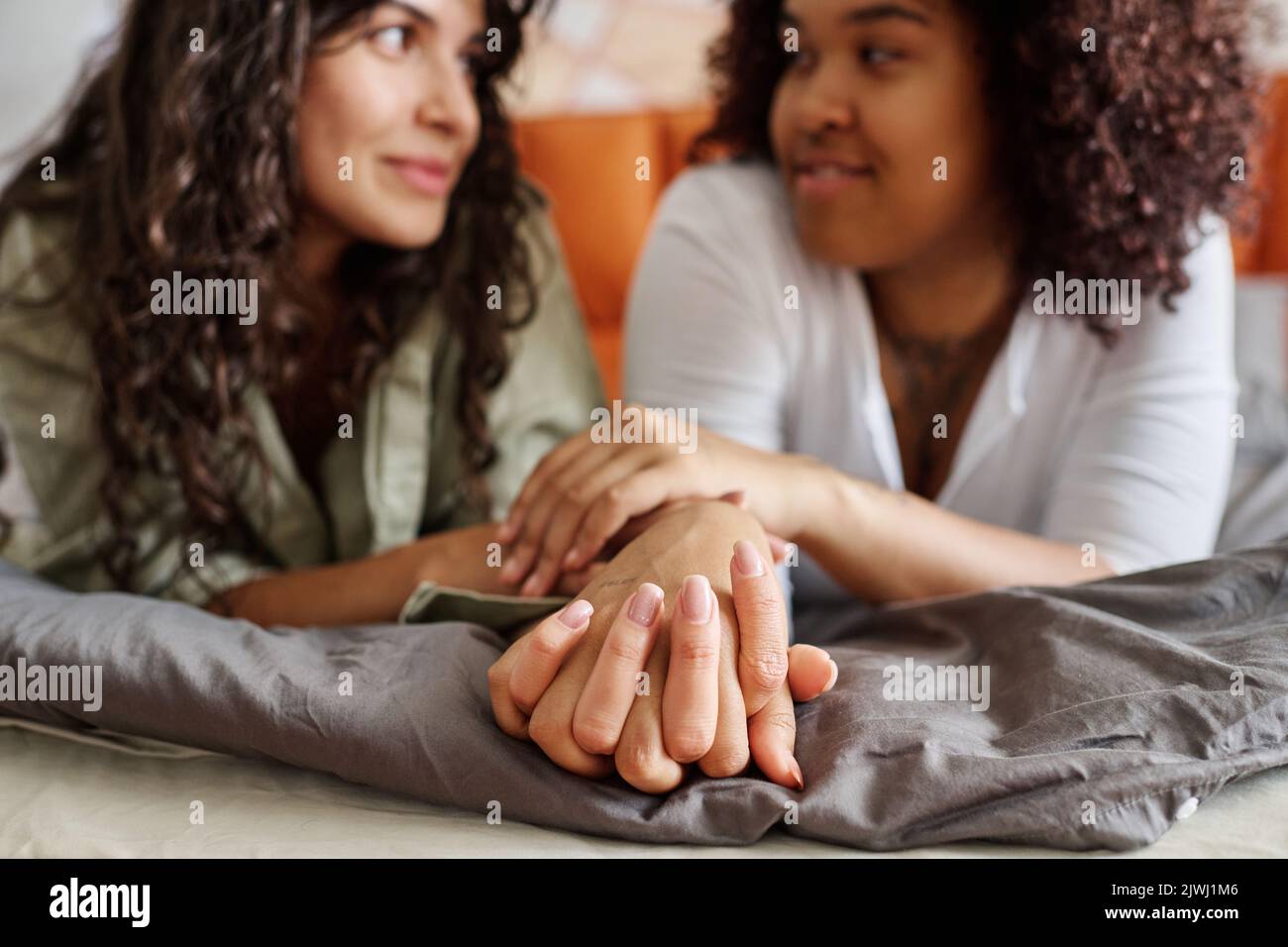 Hände von zwei jungen interkulturellen, liebevollen Freundinnen, die sich beim Entspannen im Bett in häuslicher Umgebung angusten Stockfoto