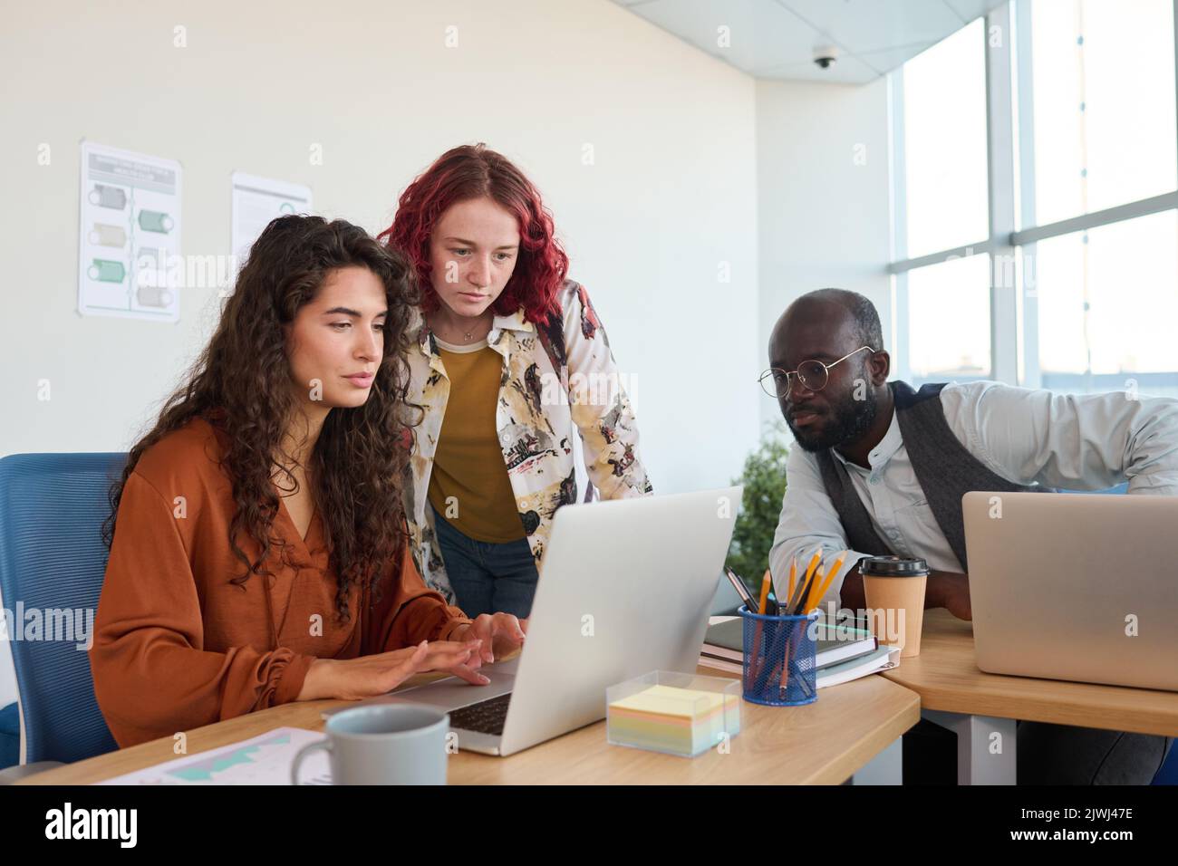 Zwei junge Geschäftsfrauen, die während der Arbeit an einem neuen Geschäftsprojekt auf den Laptop-Bildschirm blicken, während ein männlicher Kollege in der Nähe sitzt Stockfoto