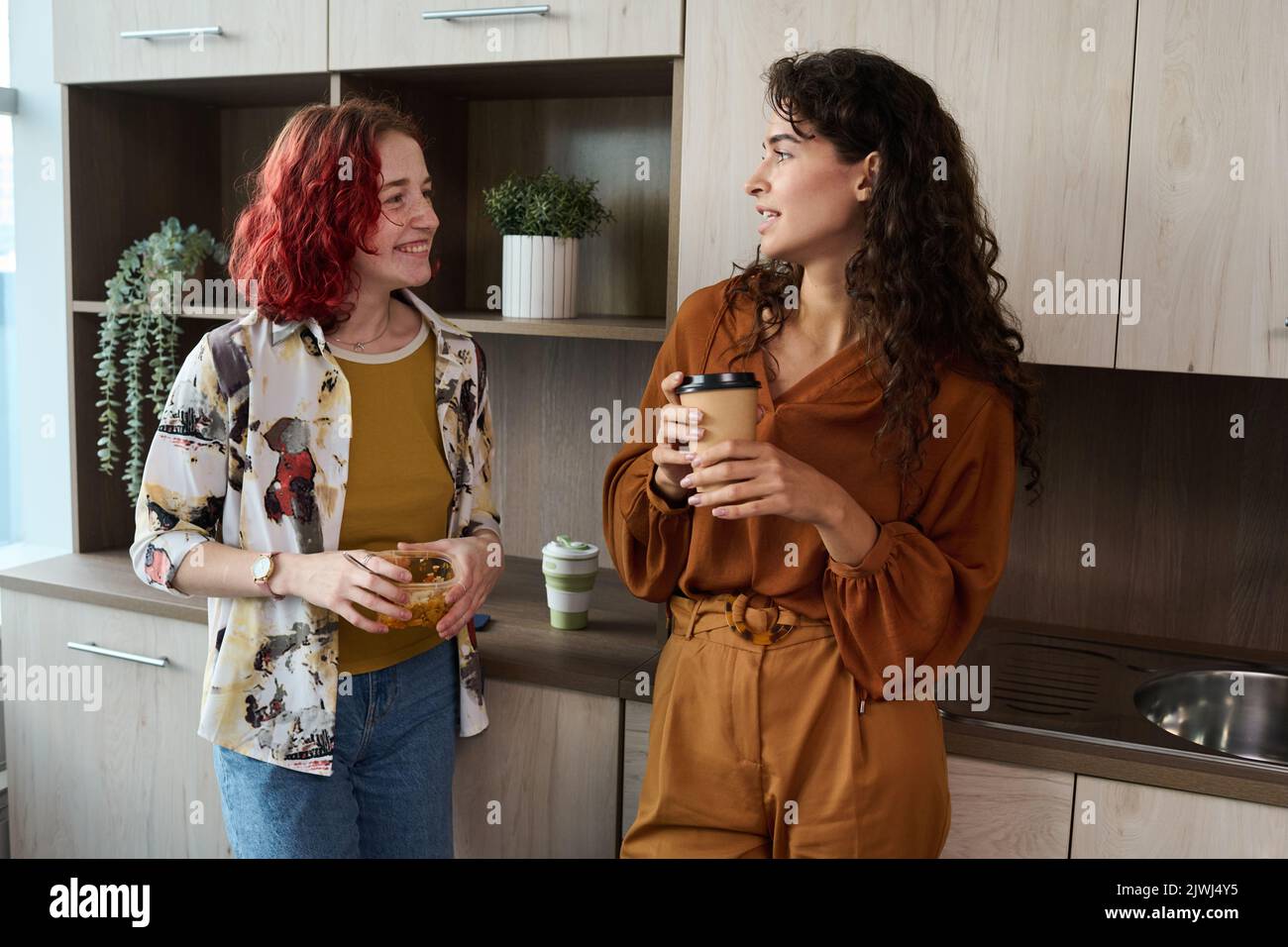 Zwei junge, elegante weibliche Angestellte in eleganter Casualwear, die Kaffee und Mittagessen trinken, während sie in der Küche stehen und in der Pause plaudern Stockfoto