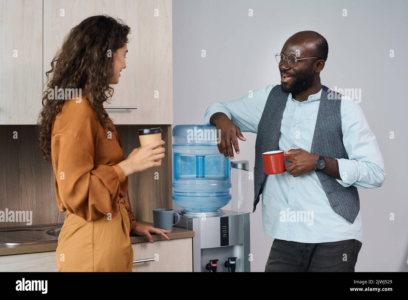 Glückliche junge interkulturelle Büroangestellte unterhalten sich und trinken in der Pause Tee und Kaffee, während sie mit Gallone am Wasserspender stehen Stockfoto