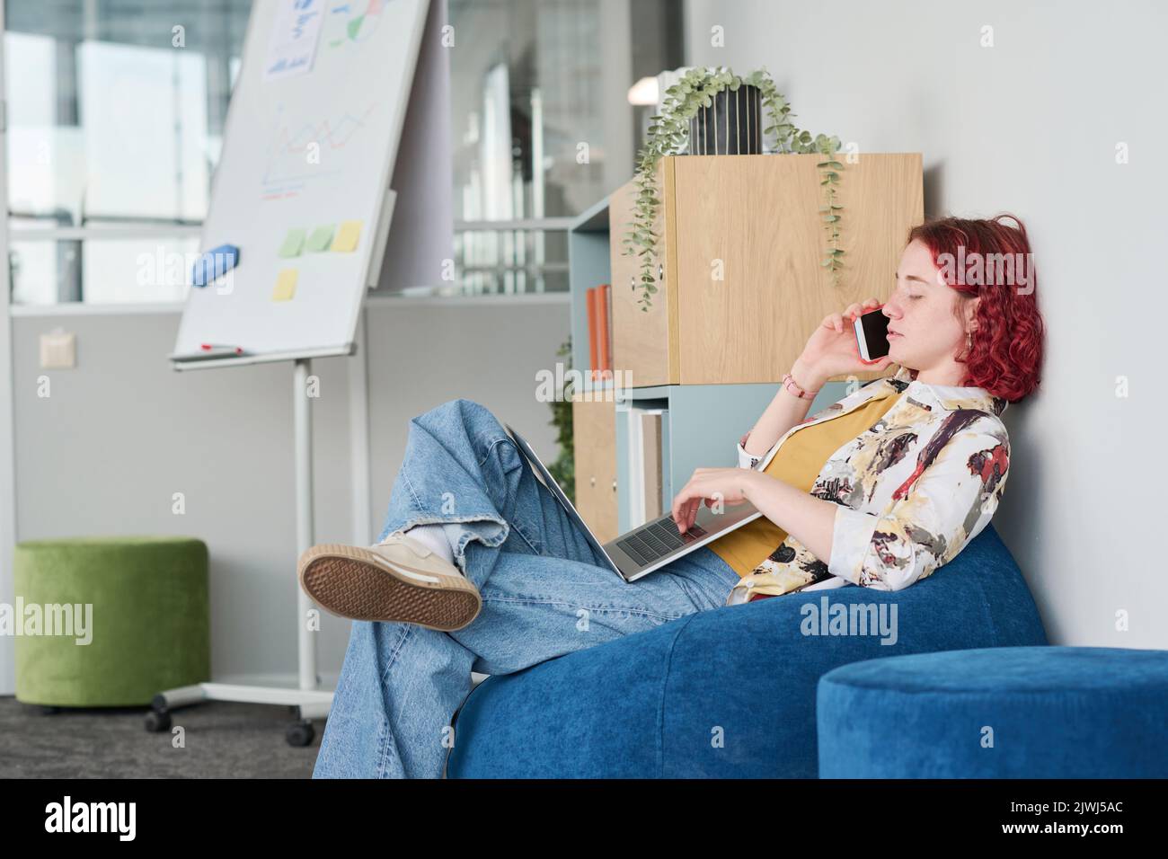 Junge, selbstbewusste Büromanagerin mit mobilen Geräten, die in einem blauen Samtsessel sitzen, auf dem Smartphone sprechen und auf dem Laptop-Tastenfeld tippen Stockfoto