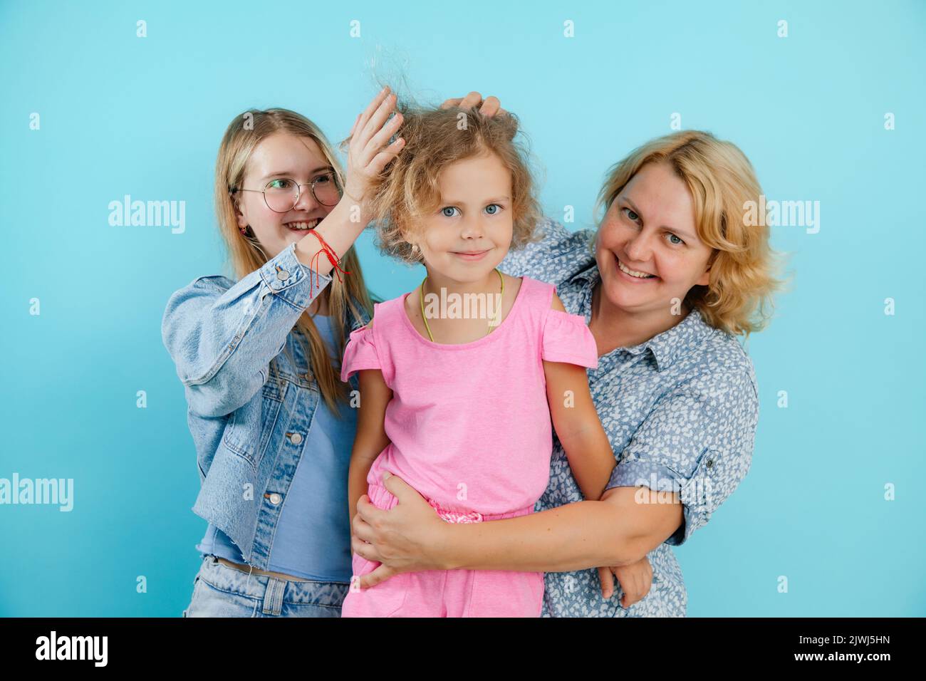 Porträt einer glücklichen Familie von drei Personen, die Spaß auf leerem blauen Hintergrund haben, freien Kopierraum. Ältere Schwester Rüschen Haare der jüngeren hübschen Schwester Stockfoto