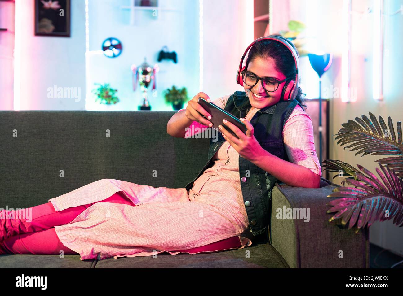 Aufgeregt Mädchen spielen Videospiel auf dem Mobiltelefon mit schnurlosem Headset, während auf dem Sofa zu Hause sitzen - Konzept der Entspannung, Hobby und Unterhaltung. Stockfoto