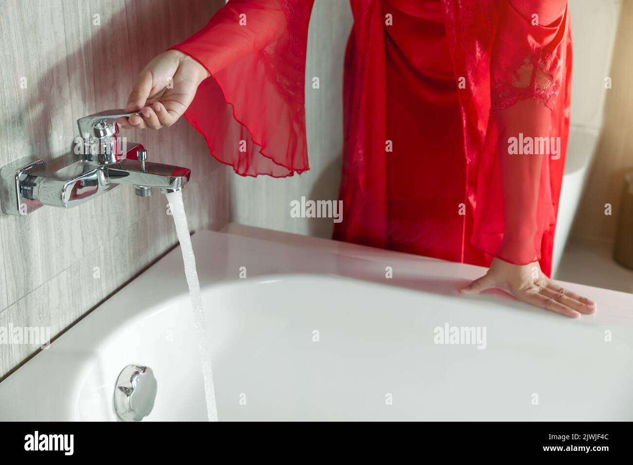 Eine Frau in einem Nachthemd aus roter Seide dreht den Wasserhahn an der Badewanne an. Frauen bereiten sich auf ein Bad vor Stockfoto