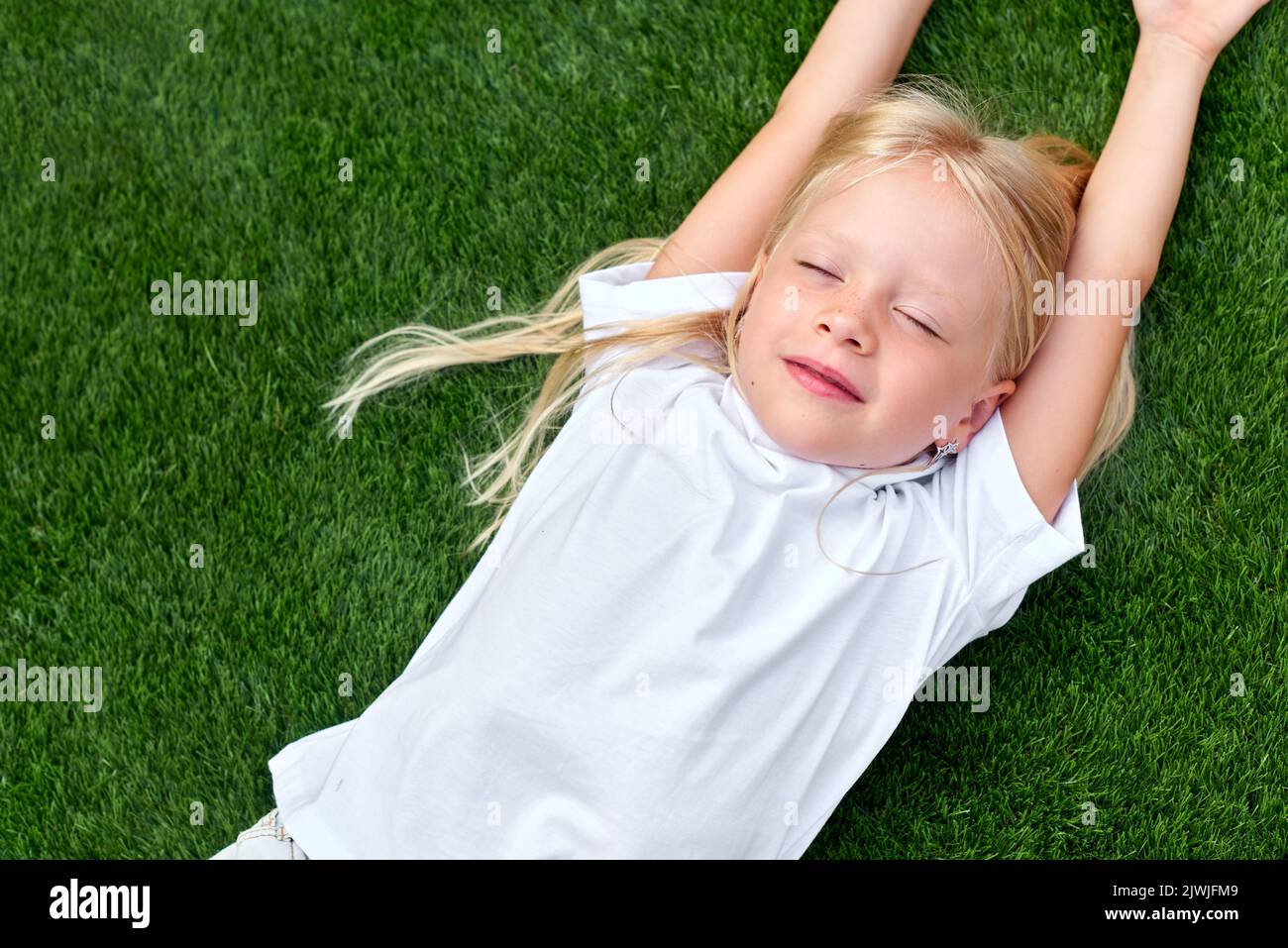 Portrait niedliches blondes Mädchen mit geschlossenen Augen auf grünem Gras liegen. Weißes T-Shirt im Lagenlook. Draufsicht. Stockfoto