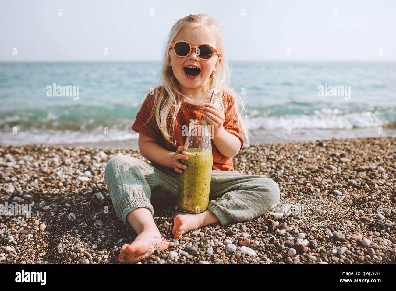 Kind trinkt Smoothie am Strand gesunder Lebensstil Sommerurlaub Kind mit wiederverwendbarer Glasflasche veganes Bio-Getränk Picknick im Freien Kind lachin Stockfoto