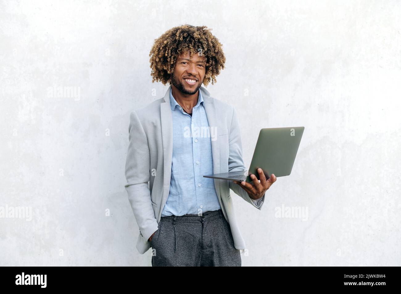 Foto des selbstbewussten intelligenten positiven erfolgreichen lockigen gemischthaarigen Geschäftsmanns, elegant gekleidet, mit offenem Laptop in der Hand, steht auf isoliertem Hintergrund, blickt auf die Kamera, lächelt freundlich Stockfoto