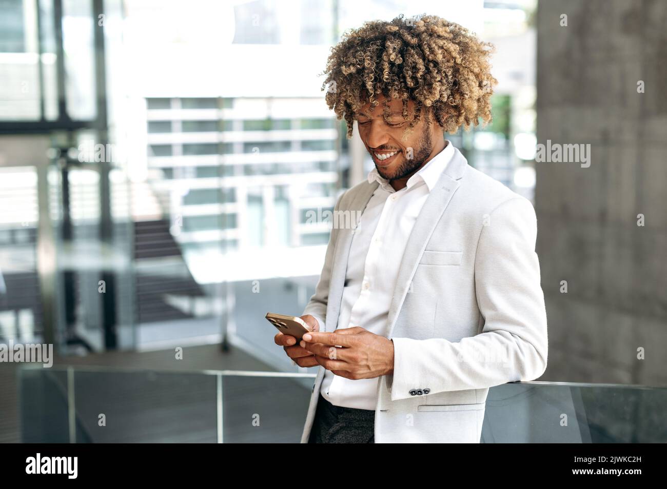 Glücklich zufrieden gemischter Renner, mit lockigen Haaren, stilvoll gekleidet, mit seinem Smartphone, während er im Freien in der Nähe des Geschäftszentrums stand, mit Freunden in sozialen Netzwerken chattete, E-Mails beantwortete und lächelte Stockfoto