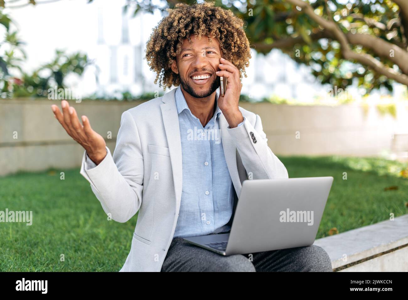 Gespräch per Handy. Busy positive erfolgreiche Mixed-Race-Business-Mann oder Top-Manager, arbeiten im Freien in Laptop, sprechen auf Handy mit Kunden oder Kollegen, schaut weg, gestikulierenden Hand Stockfoto