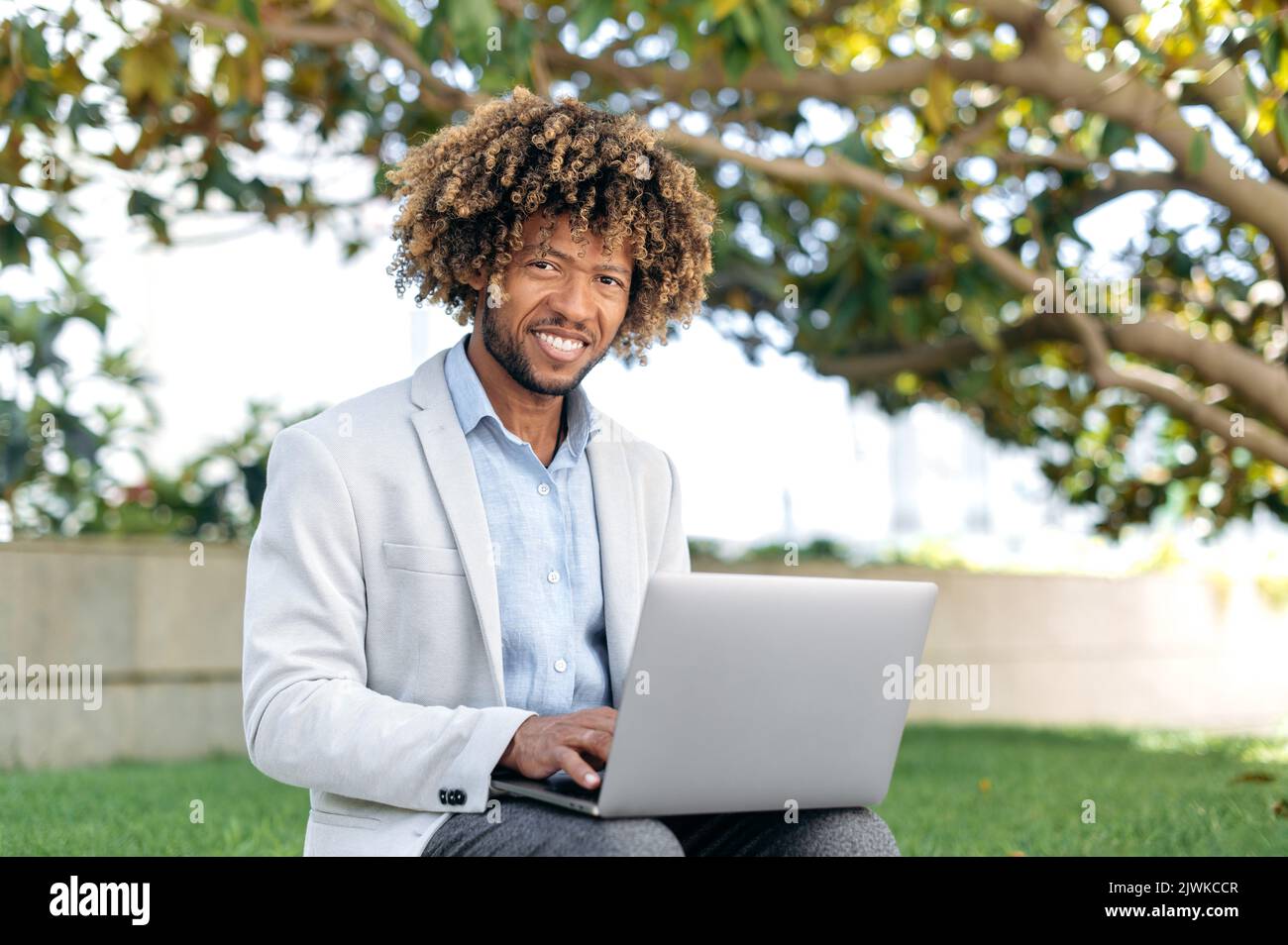 Porträt eines positiven intelligenten erfolgreichen Mixed Race Curly Mann, CEO des Unternehmens, Web-Entwickler, Content Manager, sitzen im Freien mit Laptop, arbeiten online an einem Projekt, Surfen im Internet, schaut auf die Kamera, lächeln Stockfoto