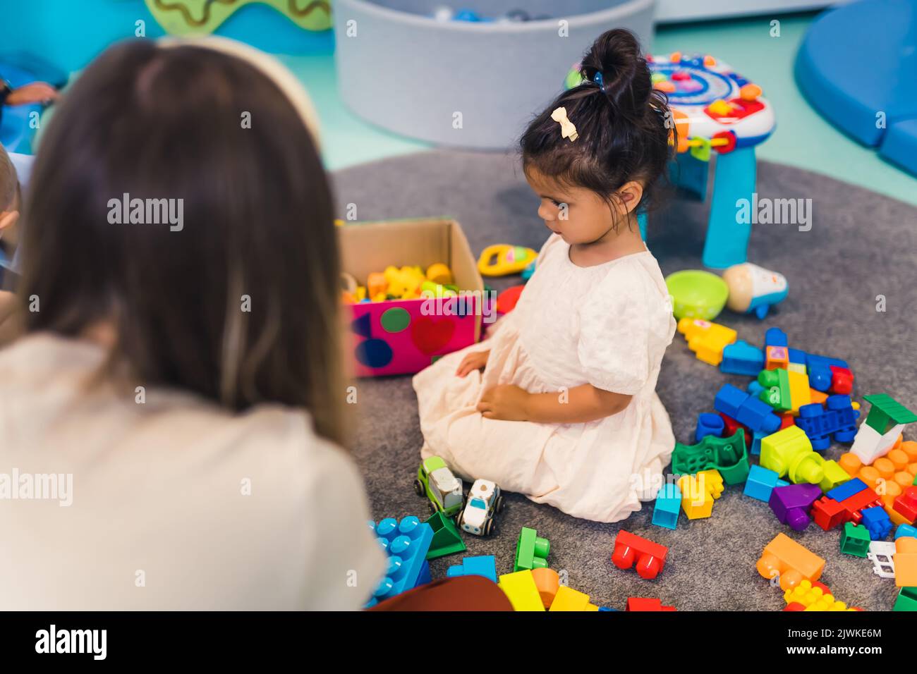 Spielerisch im Kindergarten lernen. Kleinkind kleines Mädchen und die Lehrerin spielen mit bunten Kunststoff-Spielhäusern, Bausteinen, Autos und Booten. Entwicklung von Phantasie, Kreativität, Feinmotorik und grobmotorischen Fähigkeiten. Hochwertige Fotos Stockfoto