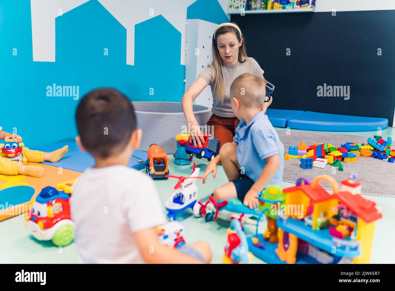 Kleinkinder und ihre Kindergärtnerin spielen mit Plastikbausteinen und farbenfrohen Autospielzeugen in einem Kinderzimmer. Frühe Entwicklung von Gehirn- und Motorik. Hochwertige Fotos Stockfoto