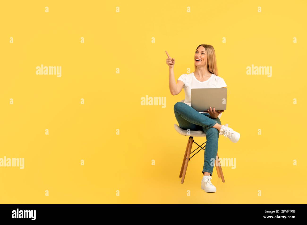 Fröhlich überrascht tausendjährige kaukasische Frau in weißem T-Shirt mit Laptop sitzt auf einem Stuhl, zeigt den Finger auf leeren Raum Stockfoto