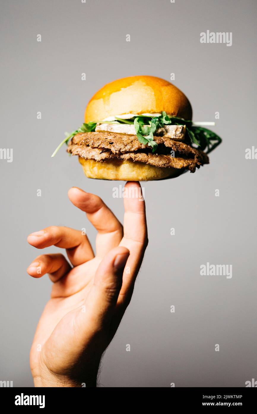 Menschliche Hand hält mit einem Finger einen Doble Burger mit altem Trockenkäse und Salat auf einem grauen Hintergrund Stockfoto