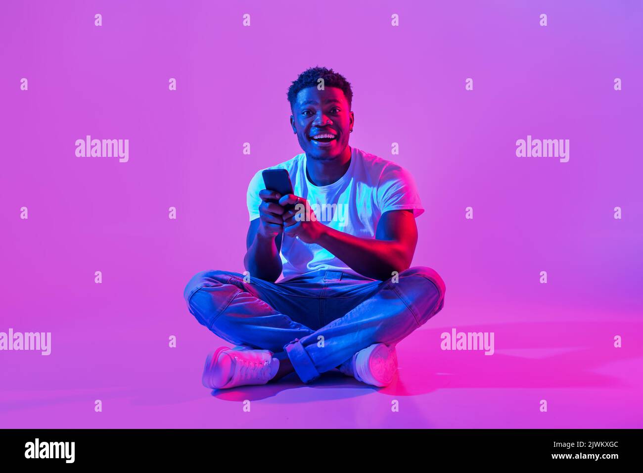 Aufgeregt junger schwarzer Kerl sitzt mit gekreuzten Beinen mit Handy, Posten in sozialen Medien, Chat im Web in Neonlicht Stockfoto