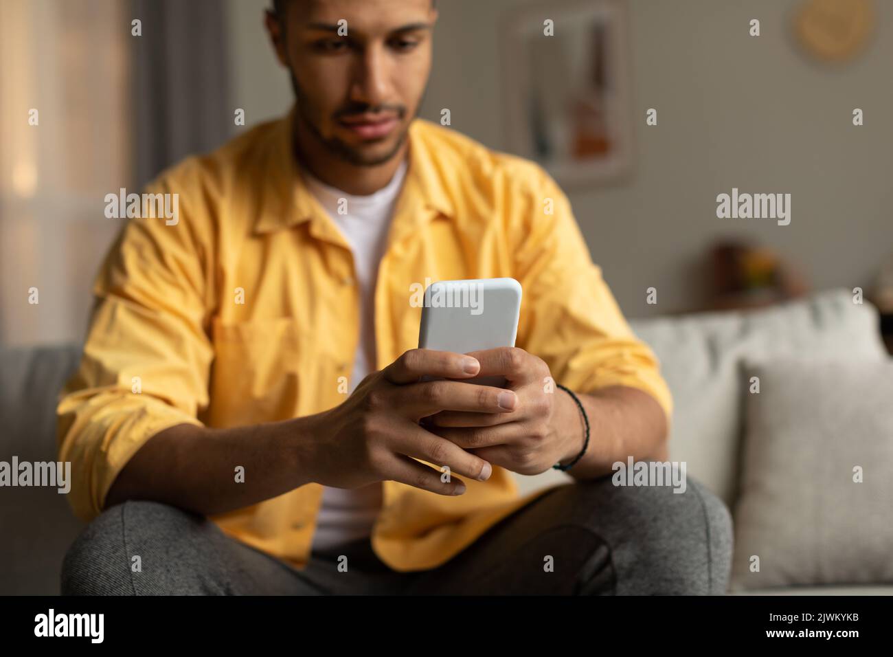 Serious junger afroamerikanischer Kerl, der mit dem Smartphone auf der Couch sitzt, online chattet und zu Hause in einem sozialen Netzwerk surft Stockfoto