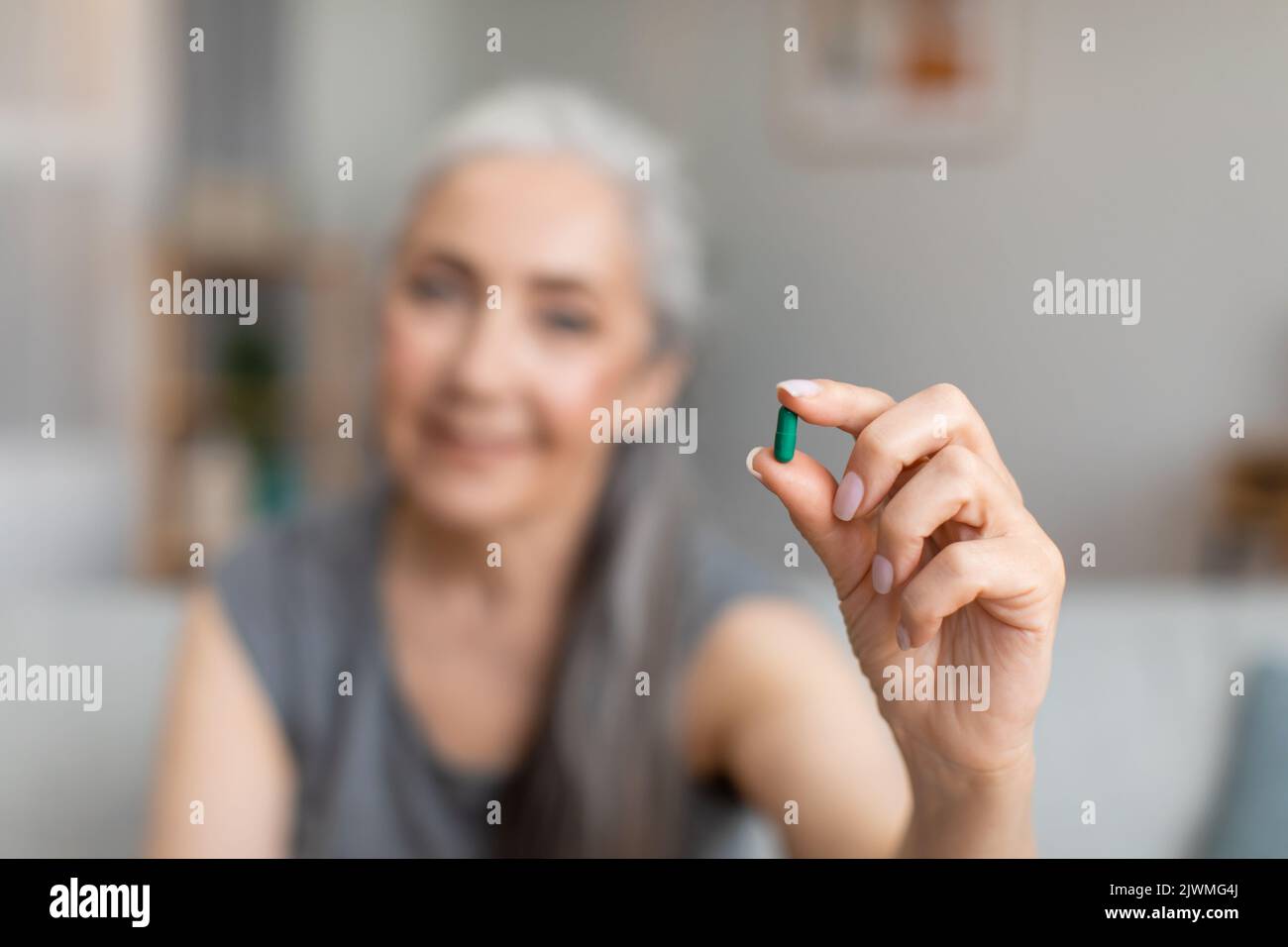 Fröhliche kaukasische ältere grauhaarige Dame hält die Pille in der Hand zur Behandlung der Krankheit im Wohnzimmer Stockfoto