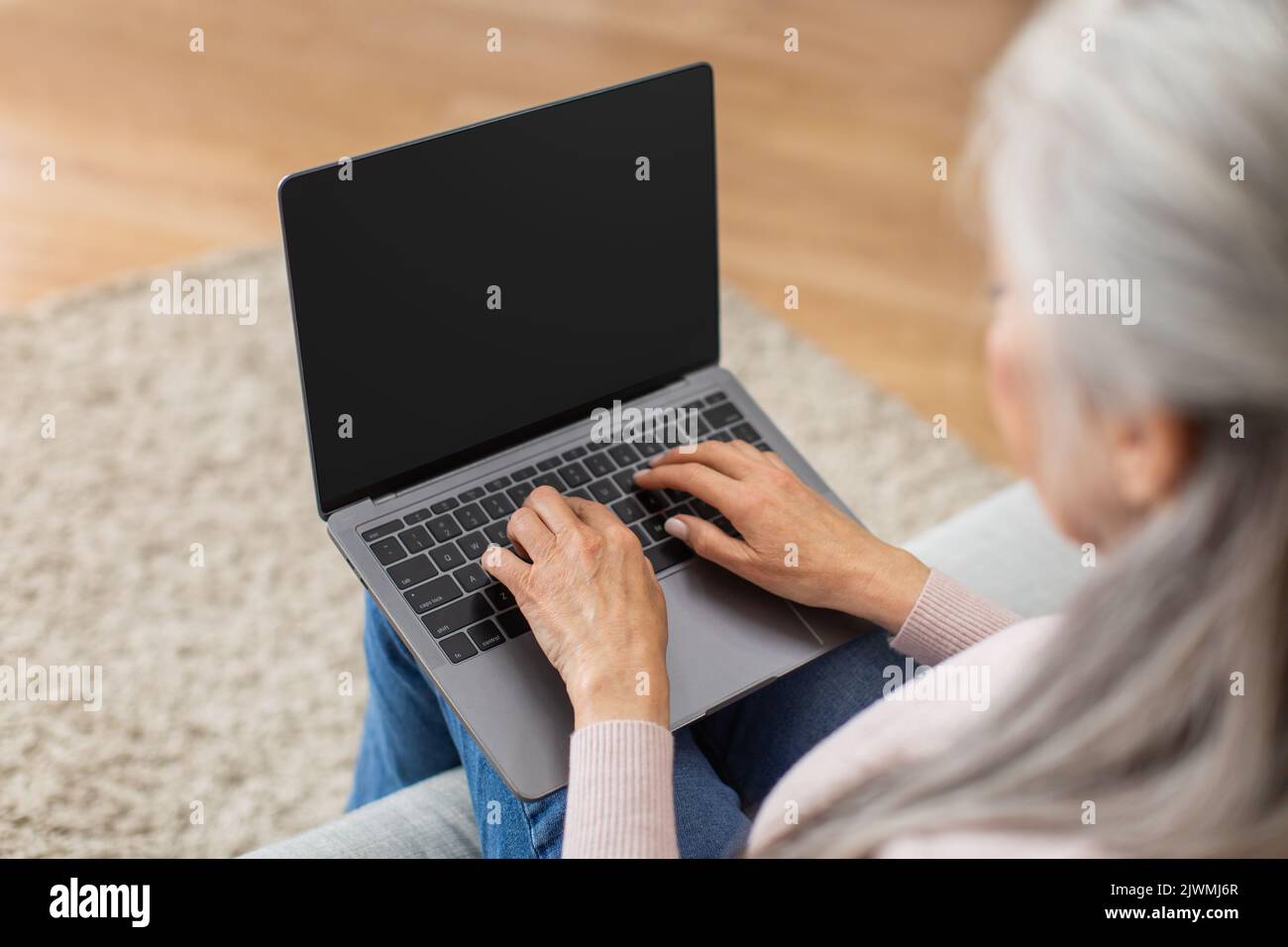 Die beschnittene kaukasische ältere grauhaarige Frau tippt auf einem Laptop mit leerem Bildschirm im Wohnzimmer Stockfoto