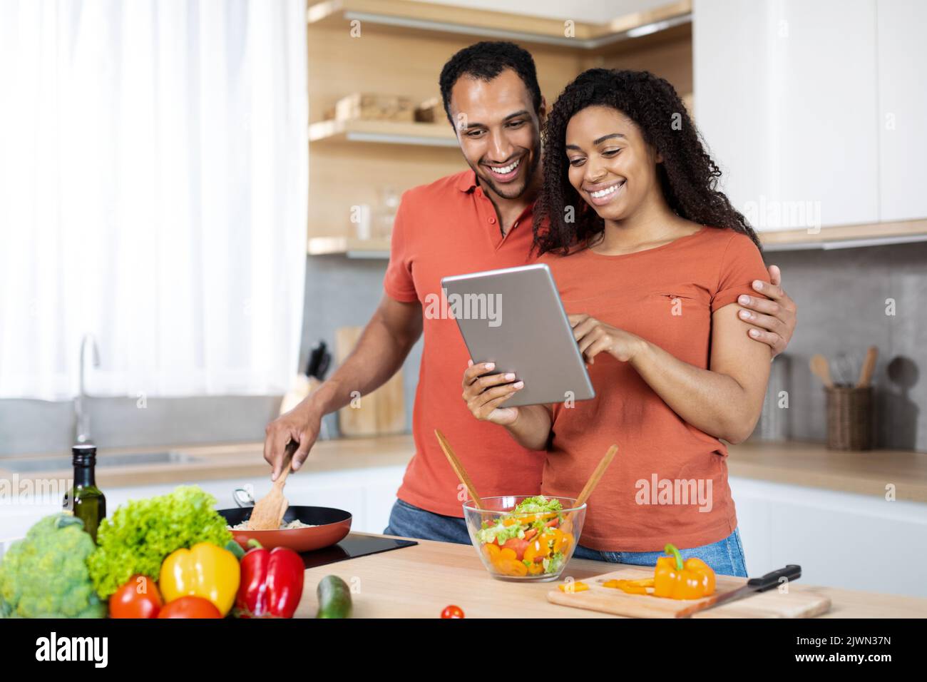 Frohes junges afroamerikanisches Paar in den gleichen T-Shirts bereiten gemeinsam hausgemachtes Essen zu, verwenden Sie Tablette Stockfoto