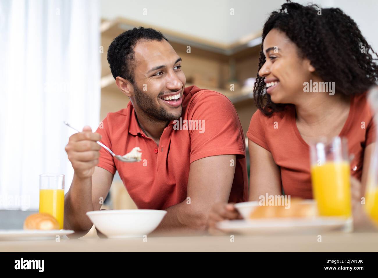 Lachende junge afroamerikanische Frau und Ehemann in gleichen T-Shirts frühstücken und genießen die Kommunikation Stockfoto