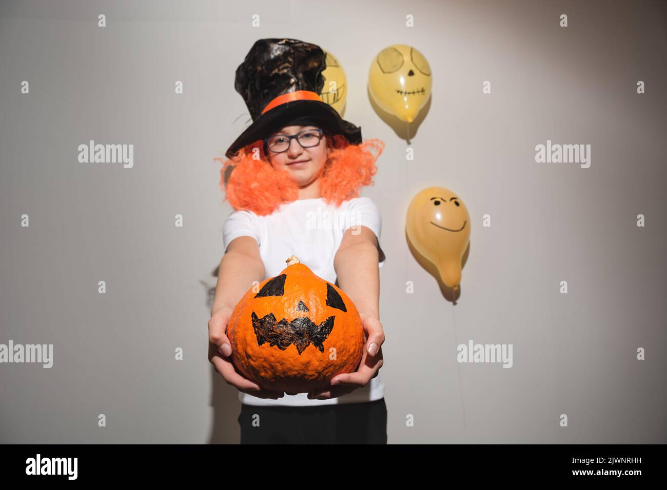 Defokussieren Sie den Vordergrund. Frohe Halloween. Kind in oranger Perücke und schwarzem Hut hält Kürbis mit gemaltem Gesicht. Das Mädchen lächelt und ist müde Stockfoto