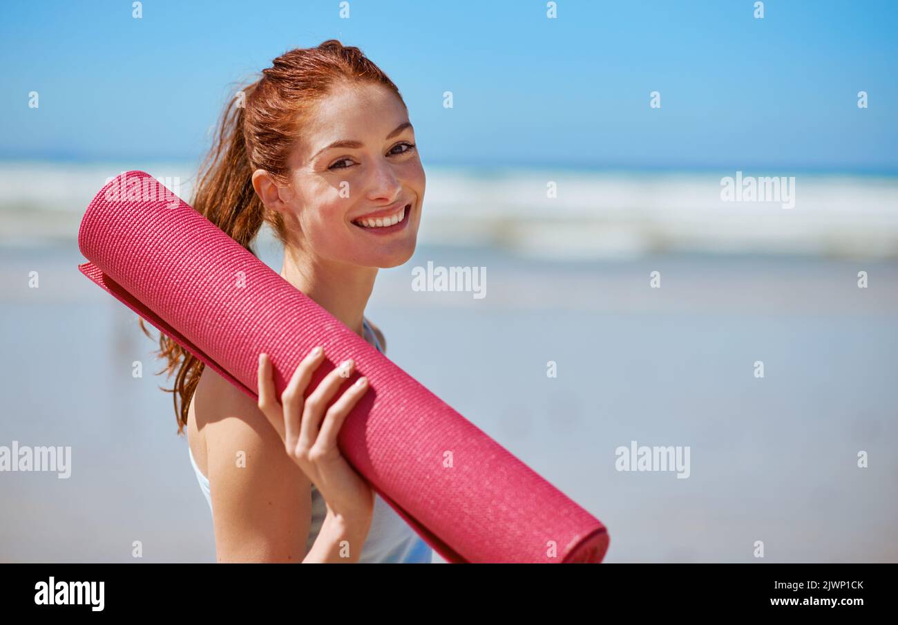 Beim Yoga geht es darum, sich auf die Matte zu zeigen. Eine junge Frau posiert mit ihrer Yogamatte am Strand. Stockfoto