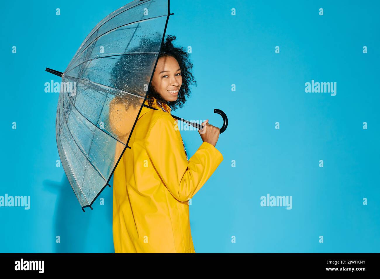 Afroamerikanische Frau in gelber wasserdichter Regenjacke mit transparentem Regenschirm isoliert auf blauem Studiohintergrund. Konzept der Herbstsaison Stockfoto
