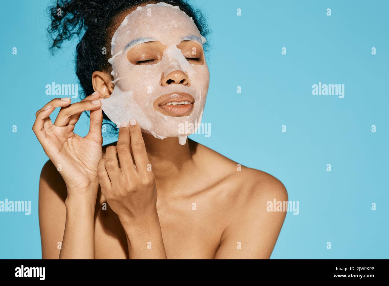 Afroamerikanische Frau wendet Kosmetik-Gewebe-Gesichtsmaske für die Feuchtigkeit spendende und feuchtigkeitsspendende Gesichtshaut auf blauem Hintergrund an. Hautpflegekonzept Stockfoto