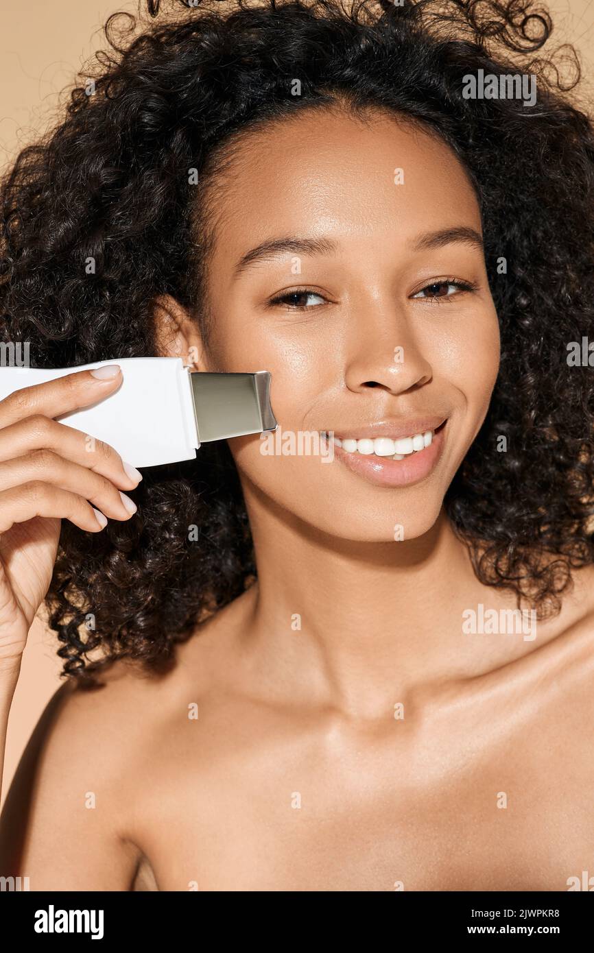 Glückliche afroamerikanische Frau mit Ultraschall Scrubber behandelt ihre Haut zum Entfernen von Mitessern und Reinigung Gesichtshaut, auf beigem Hintergrund. Ultraschall Stockfoto