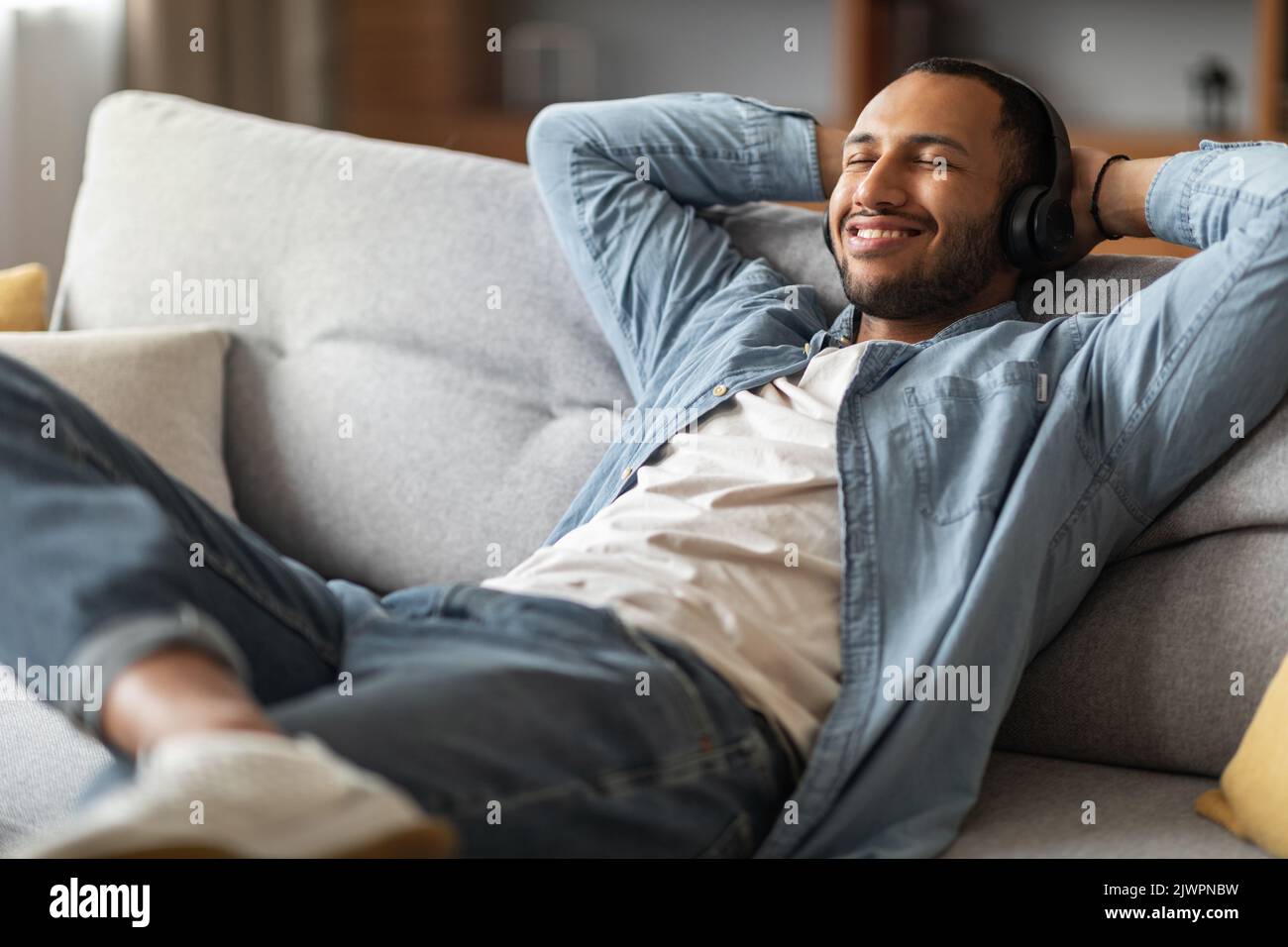 Lächelnder Afroamerikanischer Kerl, Der Musik Zuhocht, Während Er Sich Auf Der Couch Zu Hause Entspannt Stockfoto