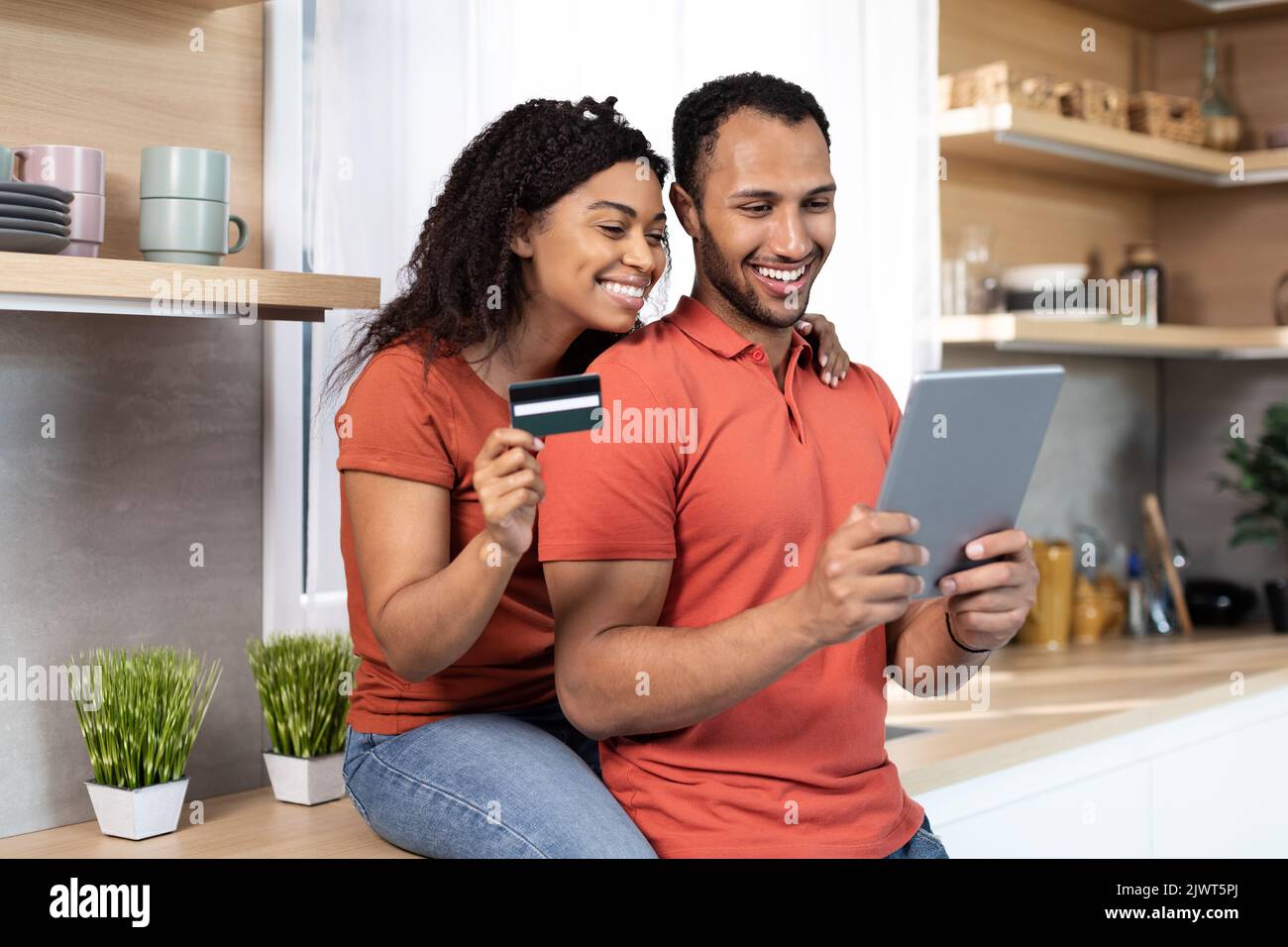 Lächelnder junger afroamerikanischer Mann und Frau in gleichen T-Shirts verwenden Kreditkarte und Tablet für Online-Shopping Stockfoto