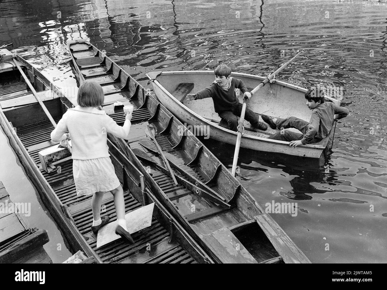 London, England, um 1967 Ein junges Mädchen tritt über festfahrende Boote, um zwei Jungen in einem Ruderboot eine Tasse Tee und eine Tüte Süßigkeiten zu übergeben. Sie sind alle Mitglieder des Piratenclubs. Der Pirate Club, ein Kinderbootclub, wurde 1966 in Gilbey's Wharf am Regents Canal in der Nähe von Camden, London, gegründet. Ihr Klubhaus war ein alter Lastkahn und eine Reihe kleiner Boote und Kanus wurden für die Kinder gespendet. Stockfoto