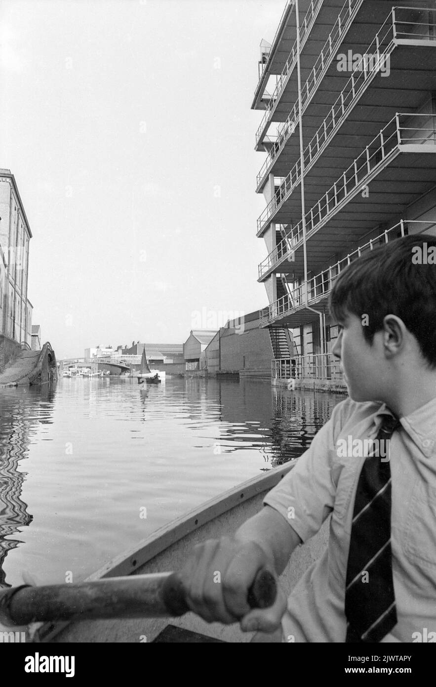 London, England, um 1967. Ein Junge in einem kleinen Boot, der am Regent’s Canal in Camden entlang rudert und rechts am Gilbey House vorbeifährt. Er ist Mitglied des Piratenclubs. Ein anderes Clubmitglied segelt in der Ferne ein Schlauchboot. Der Pirate Club, ein Kinderbootclub, wurde 1966 in Gilbey's Wharf am Regents Canal in der Nähe von Camden, London, gegründet. Ihr Klubhaus war ein alter Lastkahn und eine Reihe kleiner Boote und Kanus wurden für die Kinder gespendet. Stockfoto