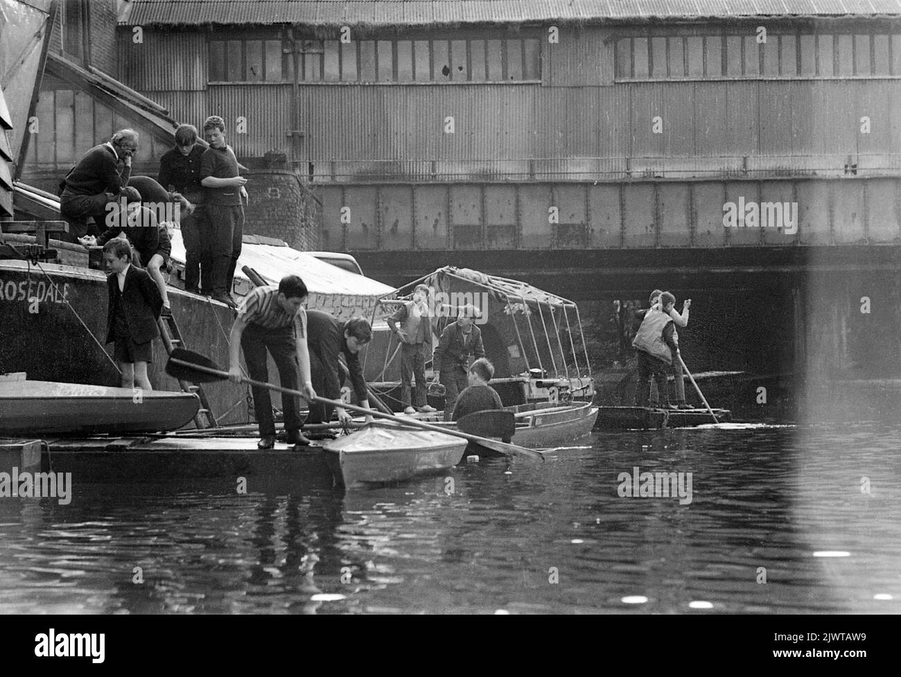 London, England, um 1967. Zwei Erwachsene Freiwillige und eine Gruppe Kinder des Piratenclubs an Bord von ‘Rosedale’, einem ausgedienten Lastkahn, der als Clubhaus diente. Andere Jungen Mann verschiedene Boote, Flöße und Kanus. Hinter ihnen befindet sich die Eisenbahnbrücke, die an die Gloucester Avenue angrenzt. Der Pirate Club, ein Kinderbootclub, wurde 1966 in Gilbey’s Wharf am Regent’s Canal in der Nähe von Camden, London, gegründet. Für die Kinder wurden eine Reihe kleiner Boote und Kanus gespendet. Stockfoto