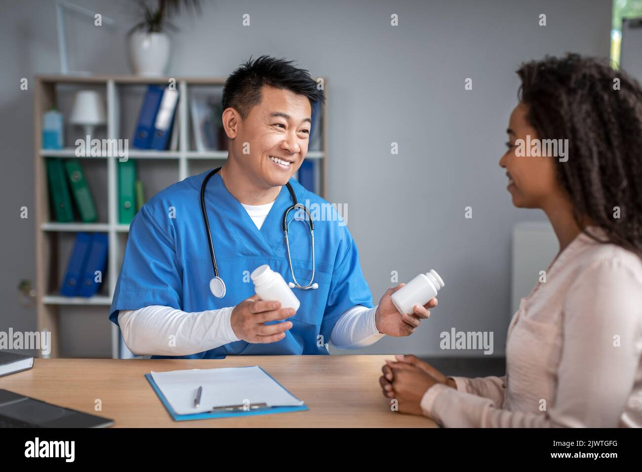 Glücklicher junger asiatischer Mann Arzt in Uniform berät schwarze Dame Patienten, bietet Gläser mit Pillen im Büro Stockfoto