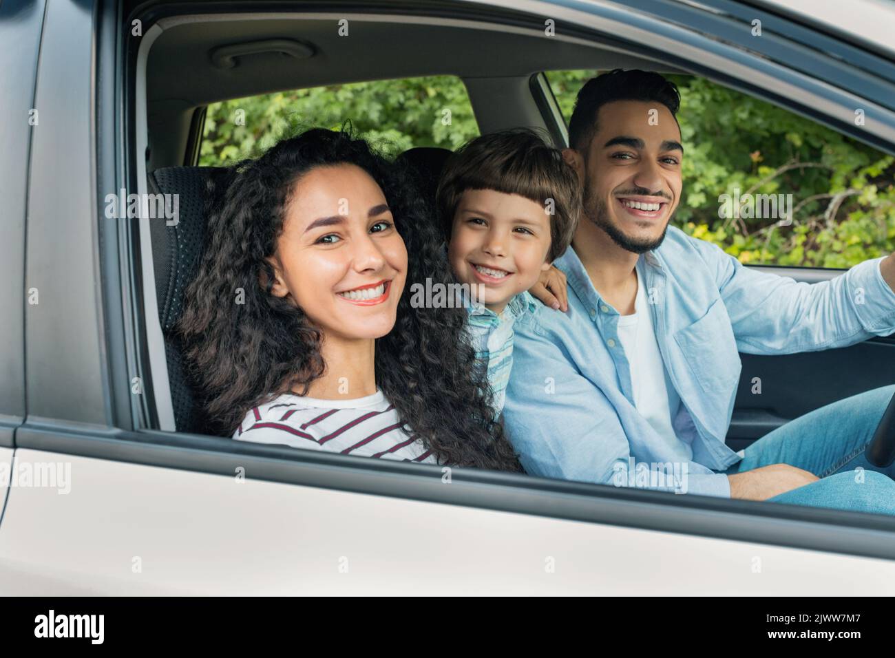 Fröhliche, fröhliche, tausendjährige arabische Mutter und Vater mit Kind fahren im Auto in den Urlaub und schauen sich die Kamera an, wenn das Fenster geöffnet ist Stockfoto