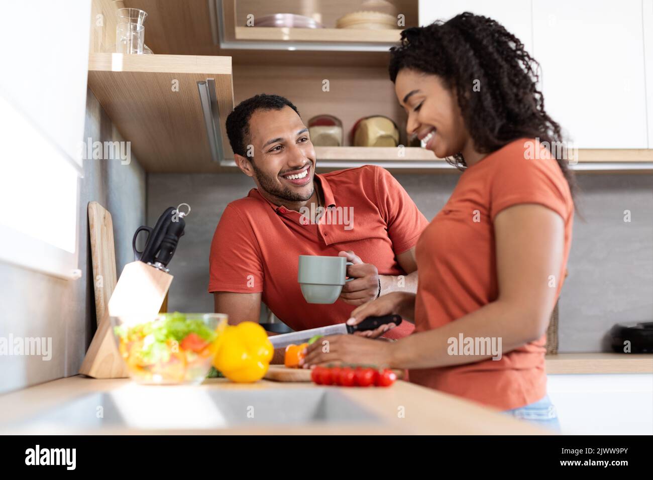 Fröhliches junges schwarzes Paar in gleichen T-Shirts, die reden, Frau bereitet Salat vor, Mann trinkt Kaffee Stockfoto